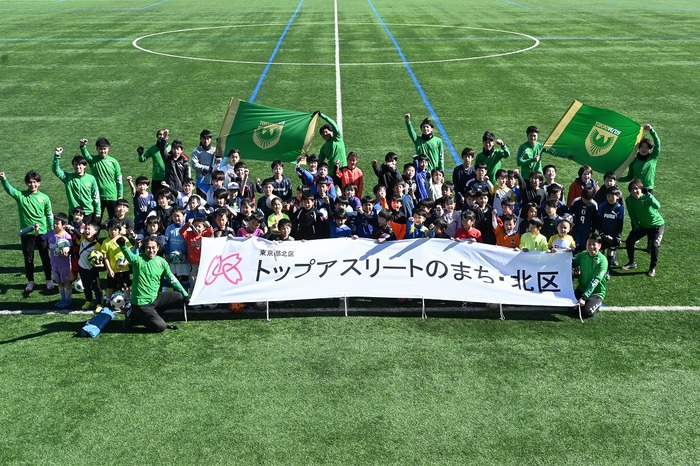 参加者・スタッフ全員で赤羽スポーツの森公園競技場のサッカー場を背景に集合写真を撮影した画像